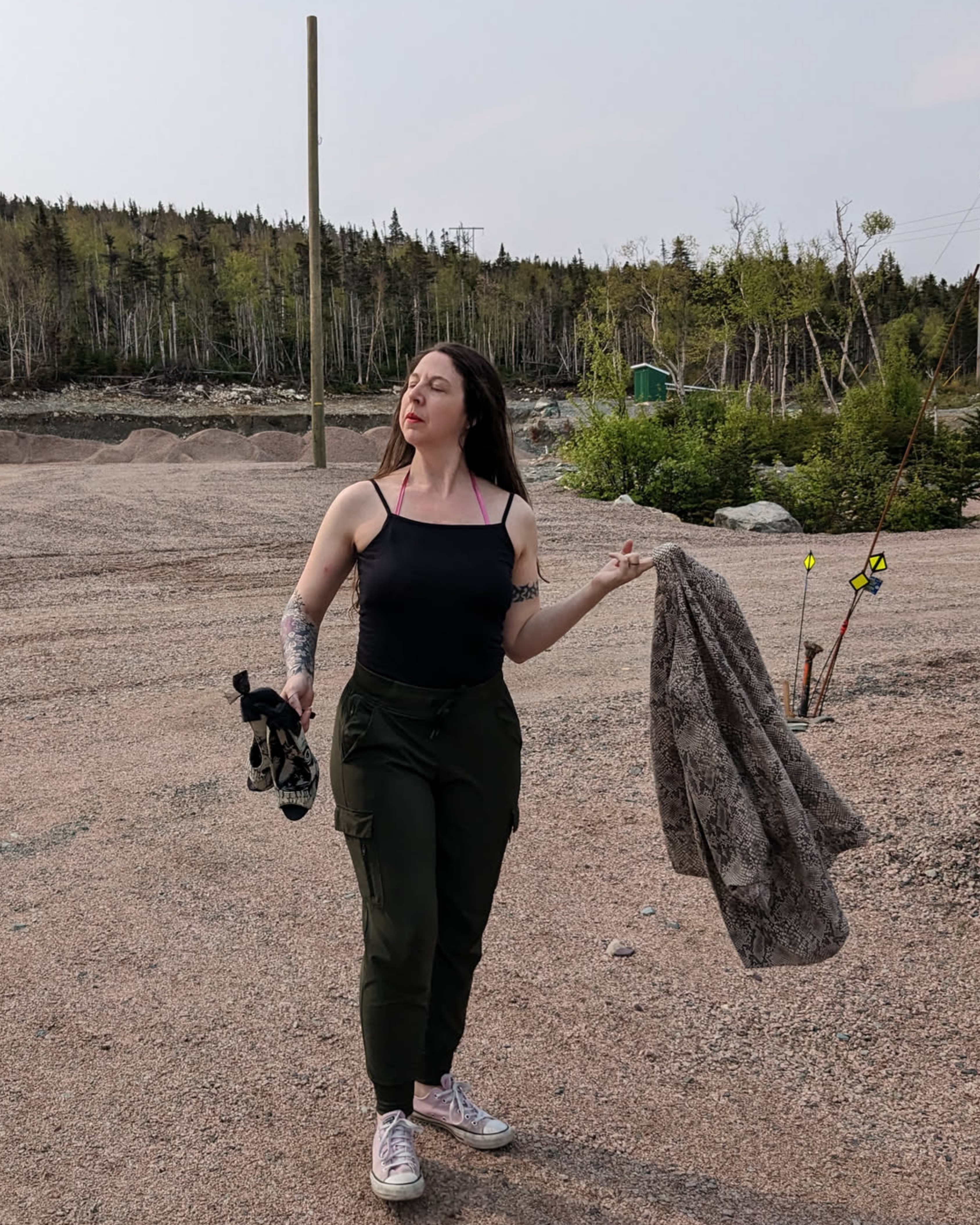 A white woman with long brown hair wearing a black tank top and track pants, pink converse sneakers, holding a pair of high heels in one hand, and a snake skin blazer in the other. She's standing in a gravel pit with a forest behind it.