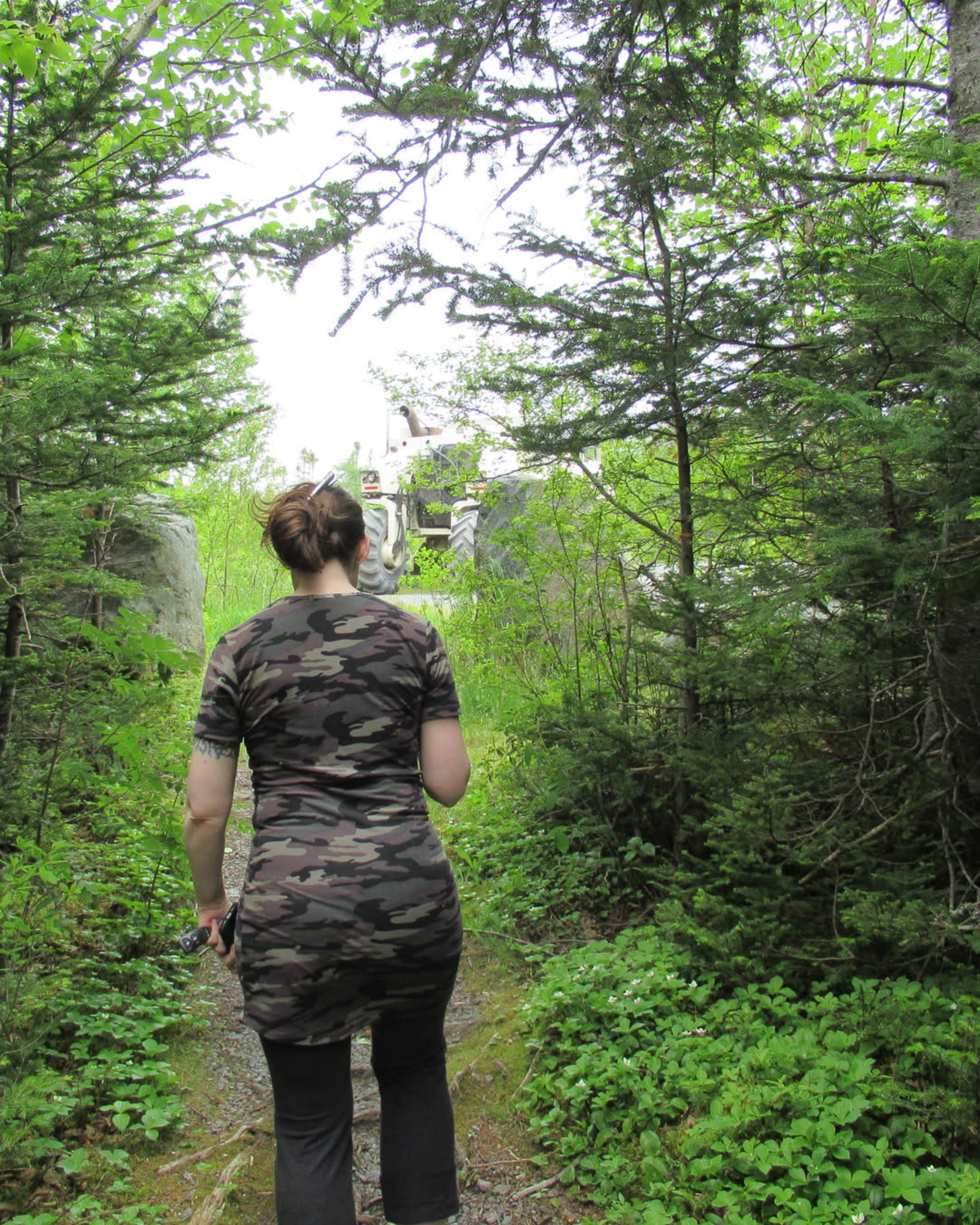 A woman in a trendy camoflauge dress shown from behind walking towards a cream coloured tractor at the end of a green forest trail. 