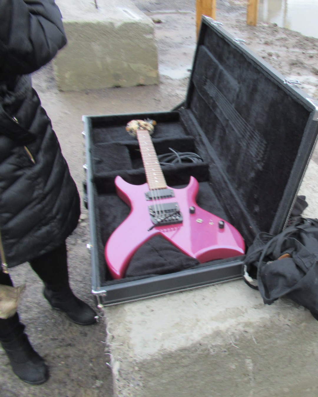 A purple BC Rich Bich electric guitar in an open case in a muddy industrial outdoor space. It's a very 80s looking pointy metal looking guitar. Kyla's black wedge boot clad feet are in the shot.