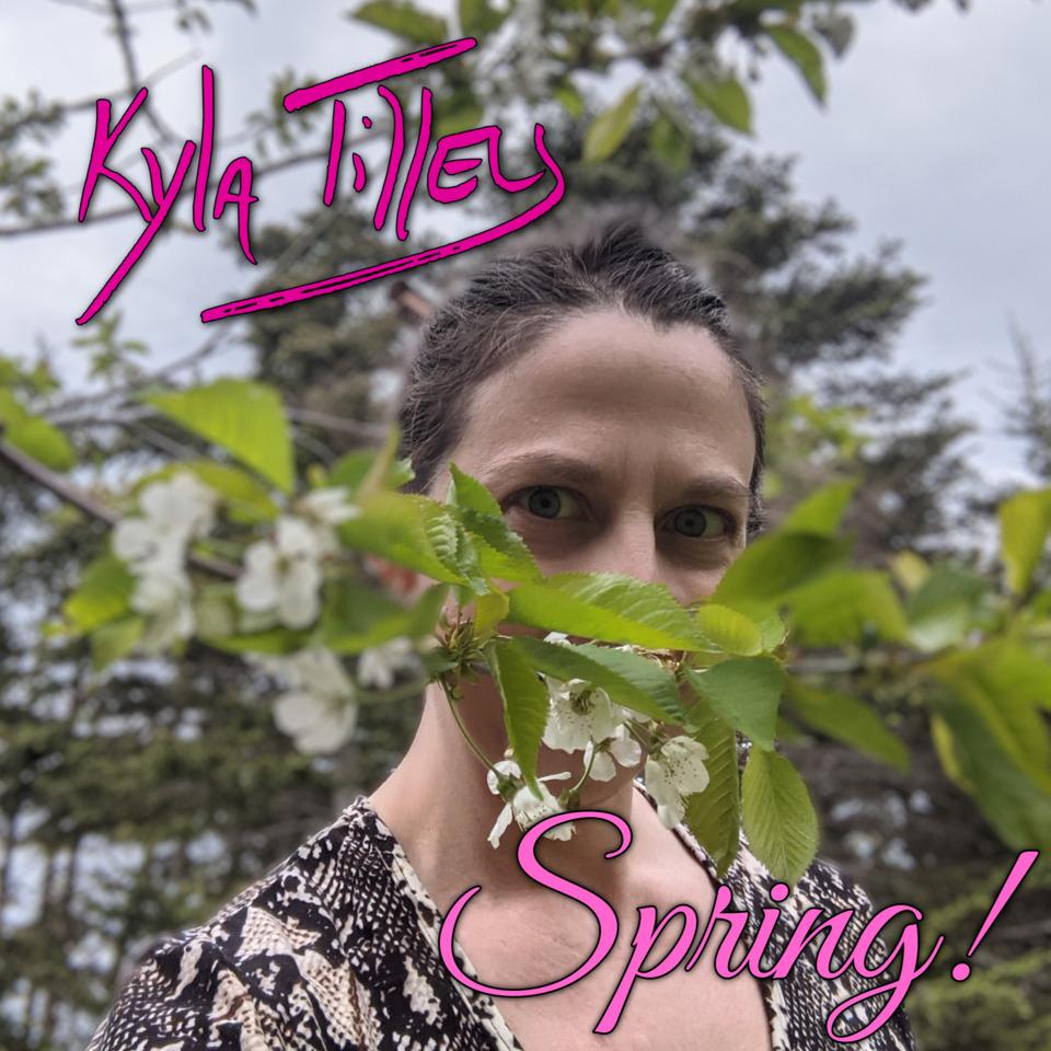 A pair of smiling blue eyes looking at you from behind a branch of a cherry tree in full bloom.  They blong to a white woman with brown hair wearing a snake print shirt.  It is a beautiful blue sky kind of day