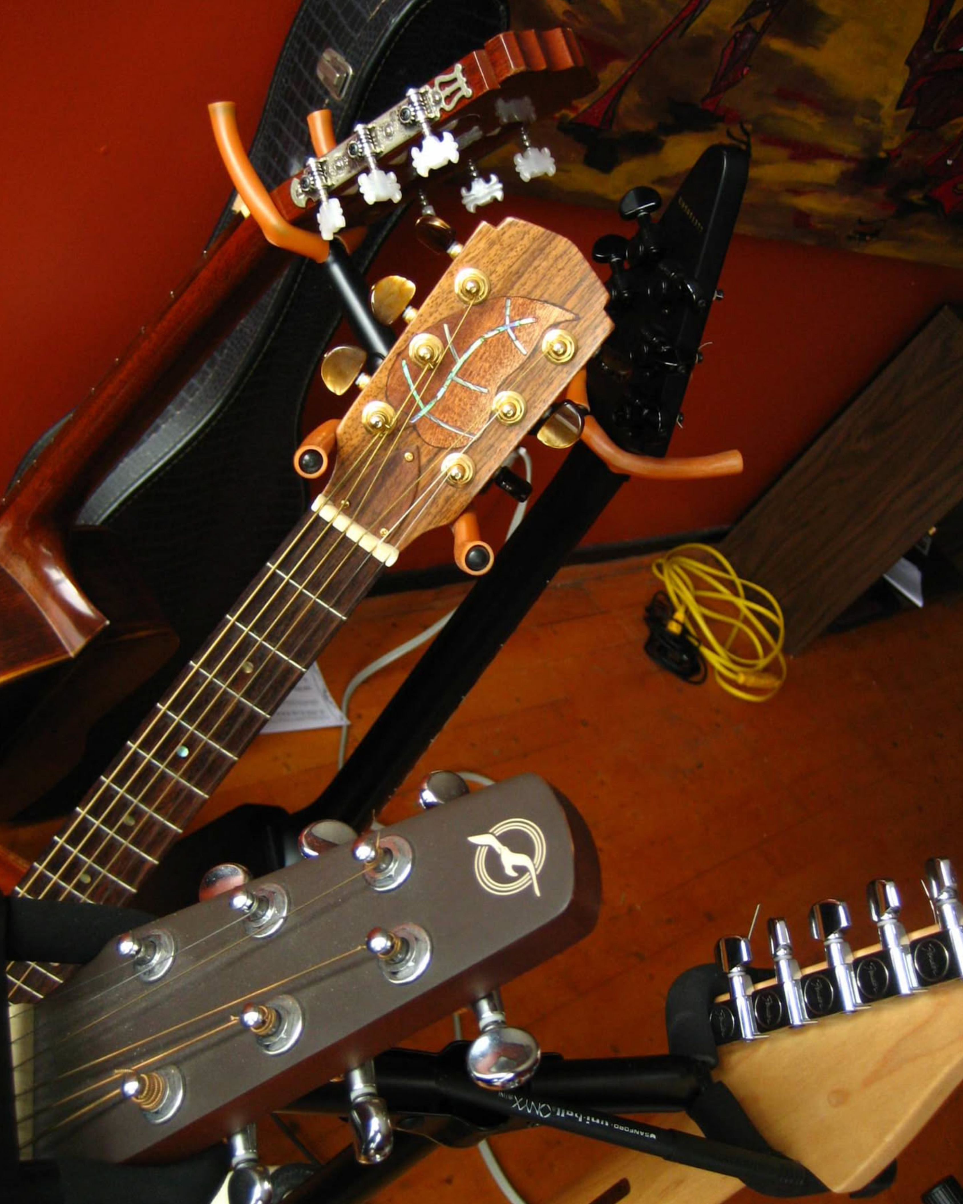 A room with dark red walls and a wooden floor. Close up on the head stocks of 5 guitars. One has a gorgeous leaf inlay on the head stock.