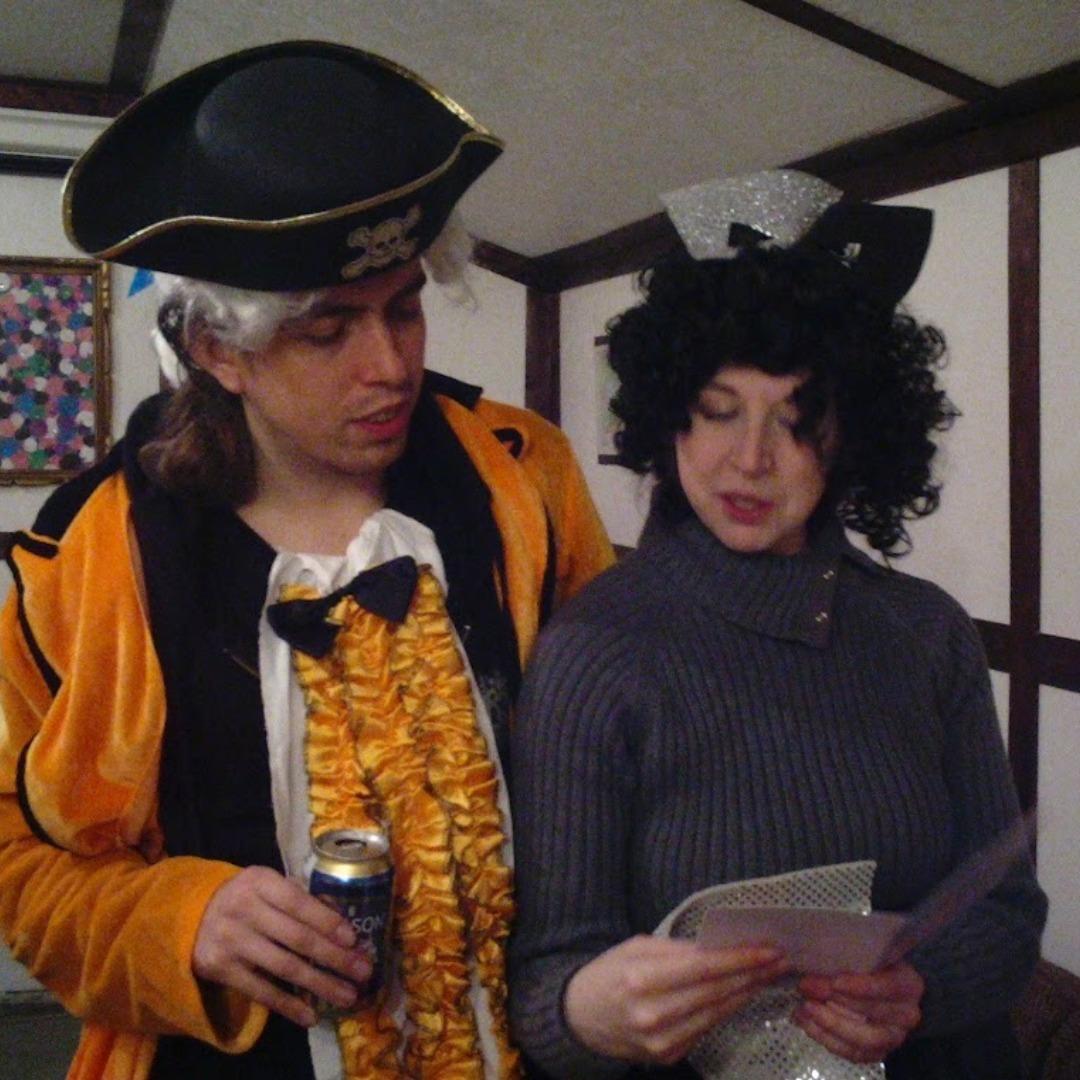  A white man in an orange 17th century aristocrat costume repleat with wig, slightly disheveled with his arm around a white woman wearing a grey sweater, a curly wig, and a head dress that looks like something you'd see in the Jetsons.  Belive it or not, these two are reading a wedding card. 