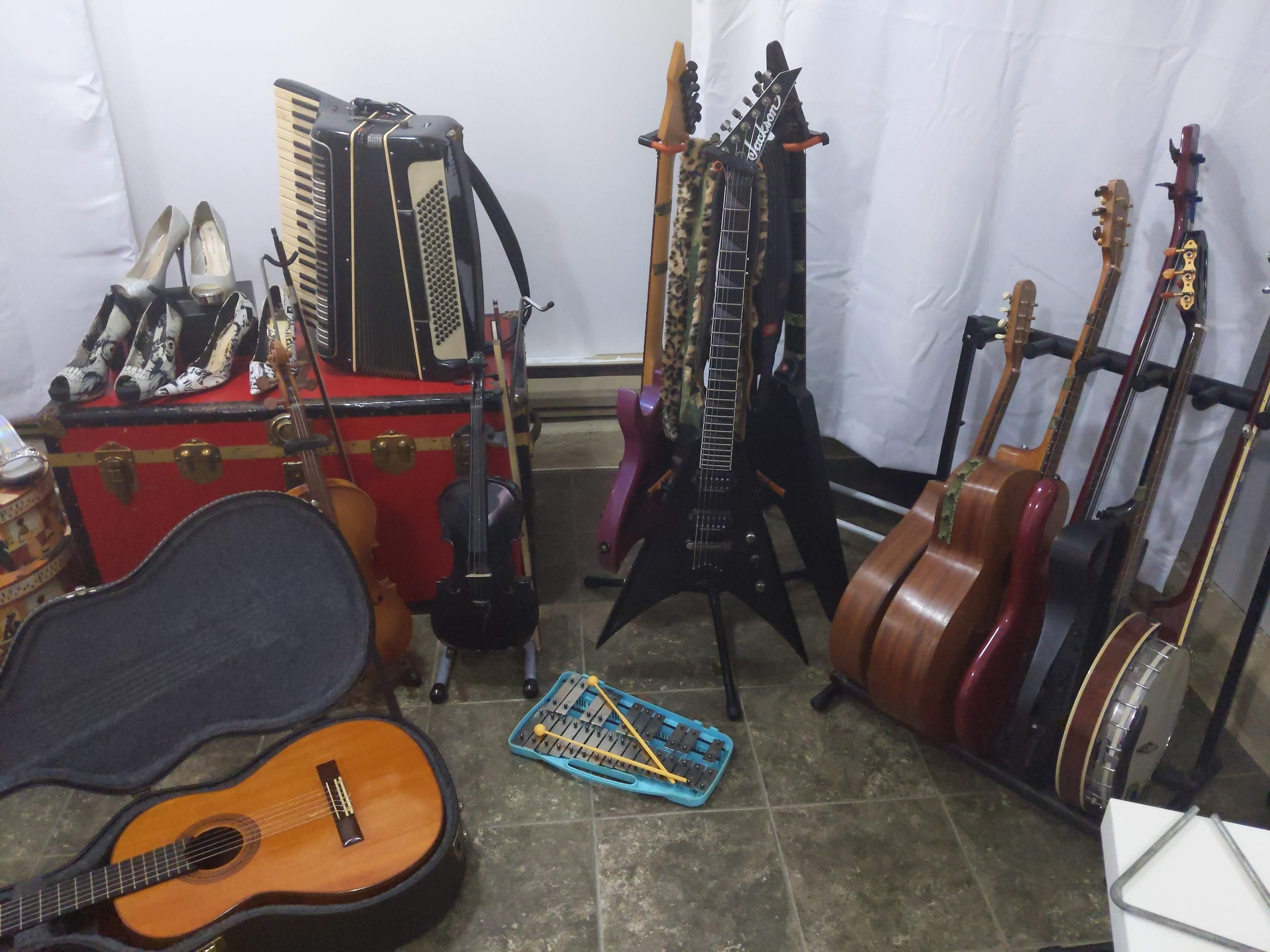 A room with a green linolium floor. On it are a glockenspiel, a black violin, a viola, a classical guitar in an open case, a red trunk with a piano accordion on top. I three guitar rack holding two black flying Vs and a...
	  
	  <a href=