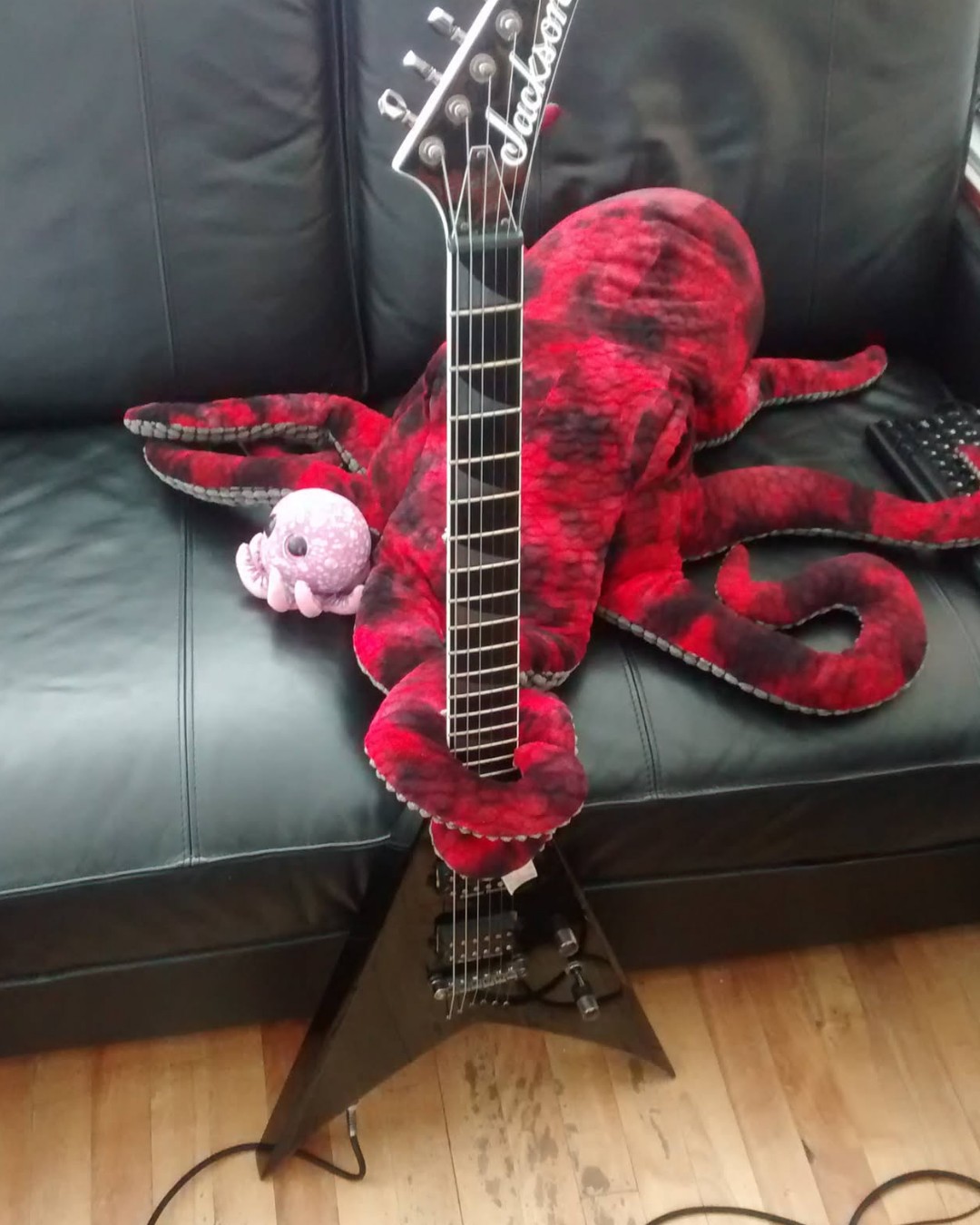 A black leather couch with a large red plush octopus with its tentacles wrapped around a shiny black Jackson Flying V electric guitar. There is a much smaller purple plush octopus snuggling with the bigger one.