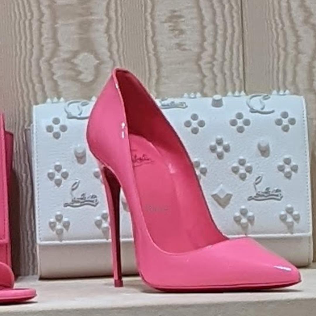 A pink high heeled shoe with a pointed toe designed by Christian Louboutin sitting on a shelf in front of a white purse. The heel is very thin, and very tall. Gosh Mr. Louboutin draws a great line! 