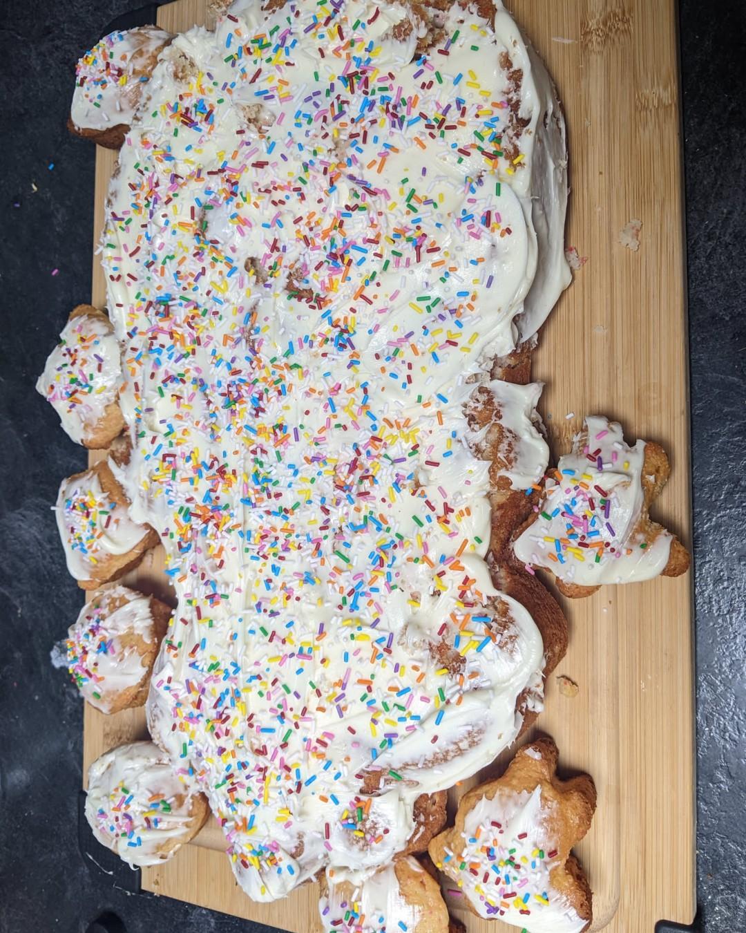  A white cake with sprinkles on a cutting board surrounded by little bug shaped cupcakes also covered in sprinkles. 