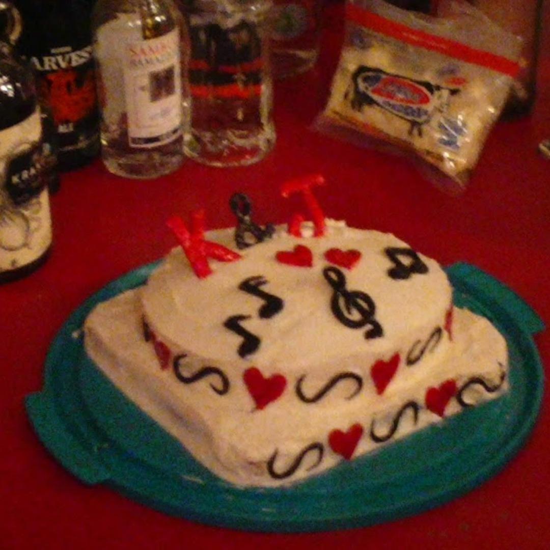 A small wedding cake decorated with hearts and music notes.  It says K & J.  It's sitting on a red table cloth, and there is a lot of booze on the table next to the cake. 