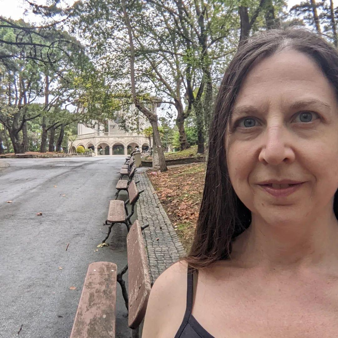 A white woman with long brown hair in a park.  There is a row of benches, and in the background a hexagonal building.