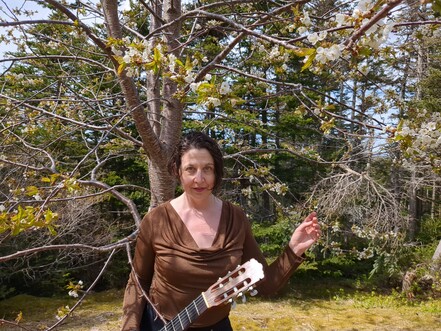 A white woman with short brown hair wearing a bronze blouse standing under a cherry tree in a very wild back yard. She's looking directly at the camera and holding a classical guitar by it's neck in her right hand, while holding her left hand up in some pseudo medieval painting position.