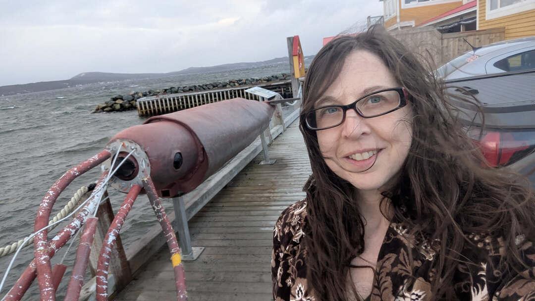 Me in my glasses wearing a brown floral blazer standing next to a sculpture of a red metal sculpture of a giant squid.  We're on a pier in Dildo Newfoundland, and you can see that it is a blustery day as my hair and the waves are being whipped around by the wind. 