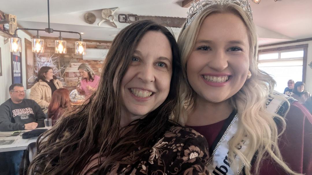 Kyla Tilley (long brown hair, brown floral blazer) snapping a selfie with Sarah Legge, the reigning Miss Newfoundland and Labrador! Sarah has long blond hair and is sporting a crown, sash, and burgundy gown.  Both have big beaming smiles.