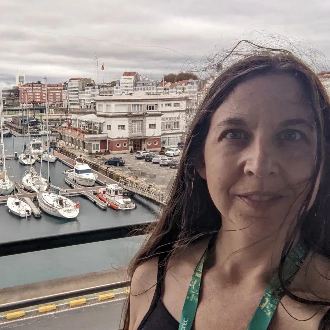 A white woman with long brown hair on a balcony with a Spanish marina in the distance.